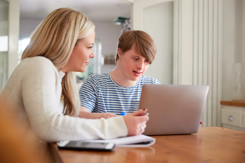 A young man and a support person sitting together at a kitchen table, looking at a laptop